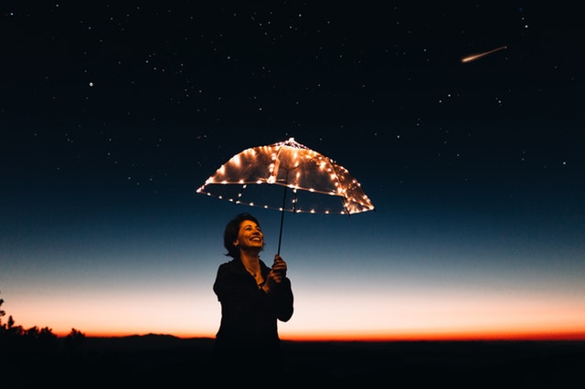 Žena stojí v tme a nad hlavou drží svietiaci dáždnik.jpg